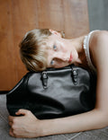 Brigitte bag in black leather, close view of the buckled straps with the model resting on it